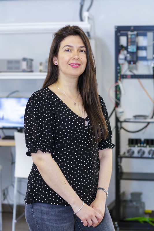 Woman in a black polka-dot blouse standing in a laboratory with equipment and cables in the background.