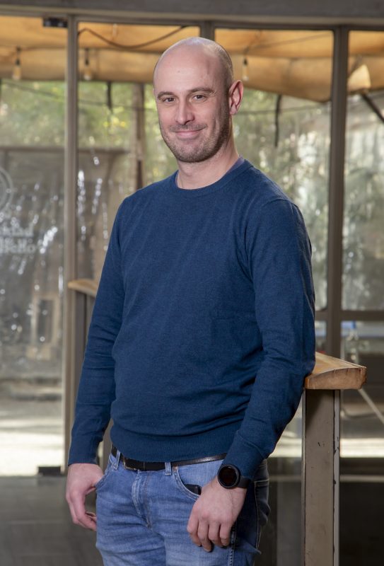 Smiling man in a blue sweater and jeans stands beside a wooden railing indoors, hands in pockets.