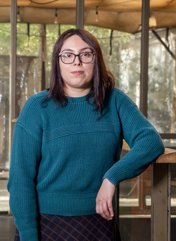 Woman with glasses and dark hair leaning on a wooden railing indoors, wearing a teal knit sweater.
