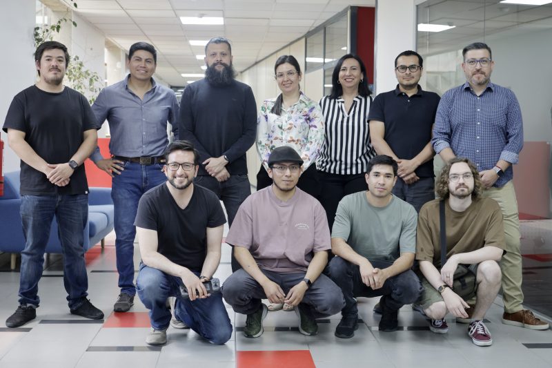 Eleven coworkers posing for a group photo in a modern office lobby with red and gray seating in the background.