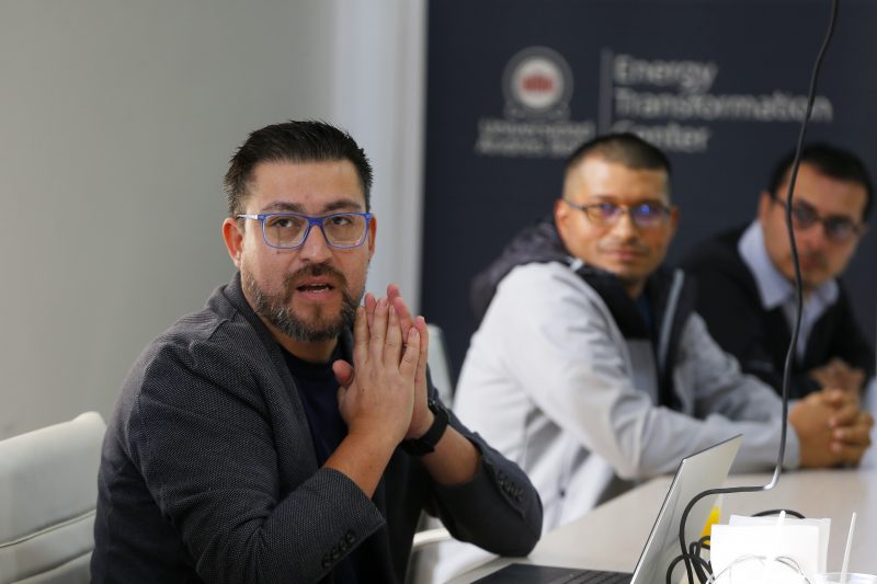 Man with blue glasses and hands clasped speaks at a panel, two listeners in the background at a conference table