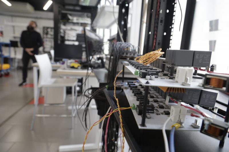 Close-up of circuit boards and wiring on a rack in a lab, with a blurred person in the background.