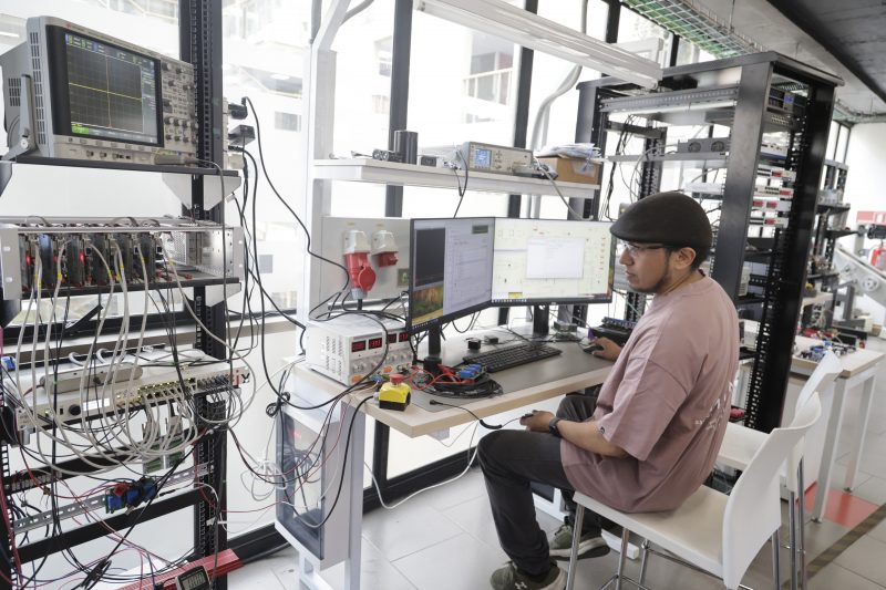 Technician in a brown shirt and cap sits at a lab bench with two computer monitors, test gear, and a rack of electronics behind him.