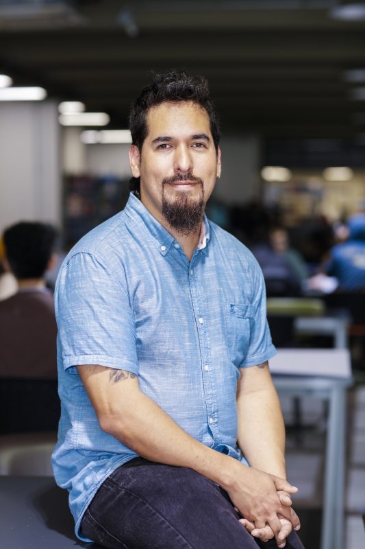 Portrait of a man in a light blue short-sleeve shirt seated in a busy coworking space, looking at the camera with a slight smile.